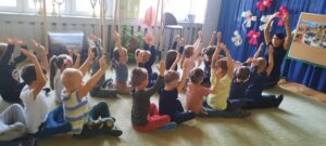 Dzieci siedzą na dywanie podzielone na dwa rzędy. Wszystkie mają podniesione obie ręce do góry.