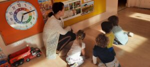 Na zdjęciu czworo dzieci siedzi na podłodze przed tablicą, do której przypięte są wizerunki zwierzą. Nauczycielka wskazuje dzieciom zdjęcie słonia.