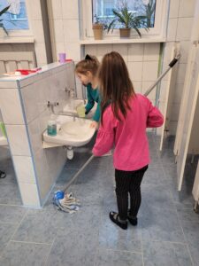 Zdjęcie przedstawia wnętrze łazienki. W łazience są dwie dziewczynki. Jedna z nich wyciera mopem podłogę, a druga myje umywalkę.