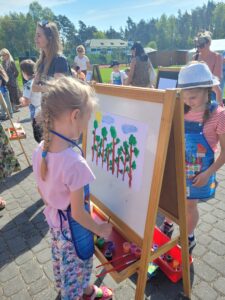 Na pierwszym planie widać dziewczynkę stojącą naprzeciw sztalugi, na której umieszczony jest karton. Dziewczynka maluje farbami las.