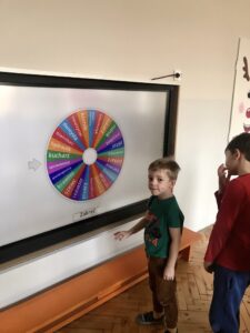 Zdjęcie przedstawia chłopca i tablicę multimedialną, na której widać kolorowe koło z wymienionymi zawodami.