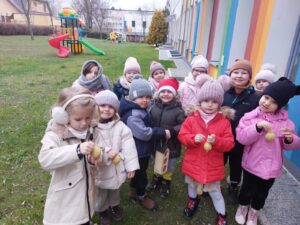Dzieci stoją w przedszkolnym ogrodzie. Trzymają w rękach karmnik i kule z ziarnami.