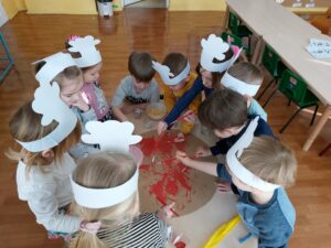 Dzieci stoja przy stoliku i nakładają na placek z papieru sos w postaci farby czerwonej 