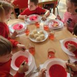 Dzieci ubrane w czerwone ubrania siedzą przy stole spożywając obiad.