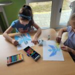 Dwie dziewczynki siedzą przy stoliku i rysują postać niebieskiego smoka