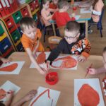 Dzieci siedzą przy stolikach i malują warzywa i owoce w kolorze pomarańczowym.