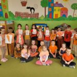 Grupa dzieci ubrana na pomarańczowo stoi obok siebie. W ręku trzymają napis ŚRODA