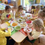 Dzieci siedzą przy stoliku i kroją owoce do sałatki owocowej.