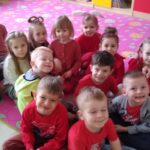 Dzieci siedzą w grupie na dywanie. Ubrane są w ubrania w kolorze czerwonym.