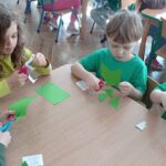 Grupa dzieci ubrana na kolor zielony siedzi przy stoliku i wycinają z zielonego papieru