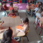 Dzieci siedzą przy stolikach i wykonują pomarańczowe prace plastyczne.