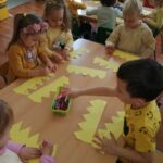 Dzieci siedzące przy stoliku wycinają koronę z żółtej kartki