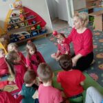 Dzieci siedzą na dywanie. Nauczycielka trzyma w ręku owoc w kolorze czerwonym.