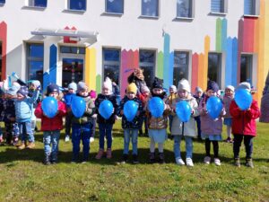 Przedszkolaki stoją przed przedszkolem. Trzymają w ręku niebieskie baloniki z napisem Światowy 